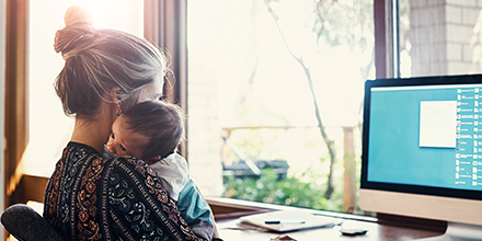 Woman holds a baby while seated at a desk, working on a computer, symbolizing career and family.