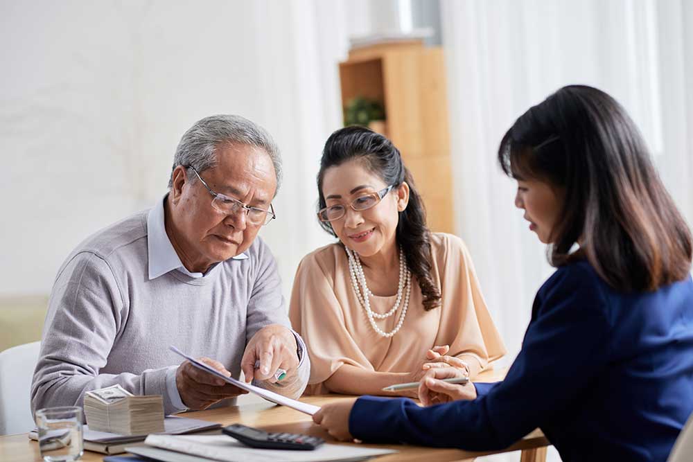 Image of an older Asian-Canadian couple sitting with their financial advisor (a woman in her 40s). The couple is facing the camera but looking at paperwork they're holding, the financial advisor is facing the couple and we see her from the back and side.